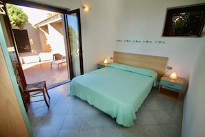 A bedroom with a bed, two bedside tables, a chair, and a sliding glass door leading to an outdoor area with a patio and a wooden chair. - Three-room villa with sea view of approximately 55 m2 furnished in a simple and essential way consisting of a living room with double sofa bed, kitchenette equipped with gas stove.A double bedroom with wardrobe and another bedroom with two single beds, b (Trinita' d'Agultu e Vignola)