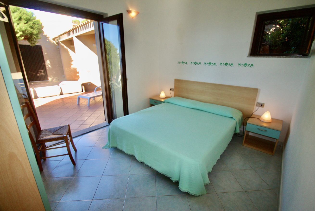 Un dormitorio con una cama, dos mesitas de noche, una silla y un ventanal corredero que da a un espacio exterior con un patio y una silla de madera.
