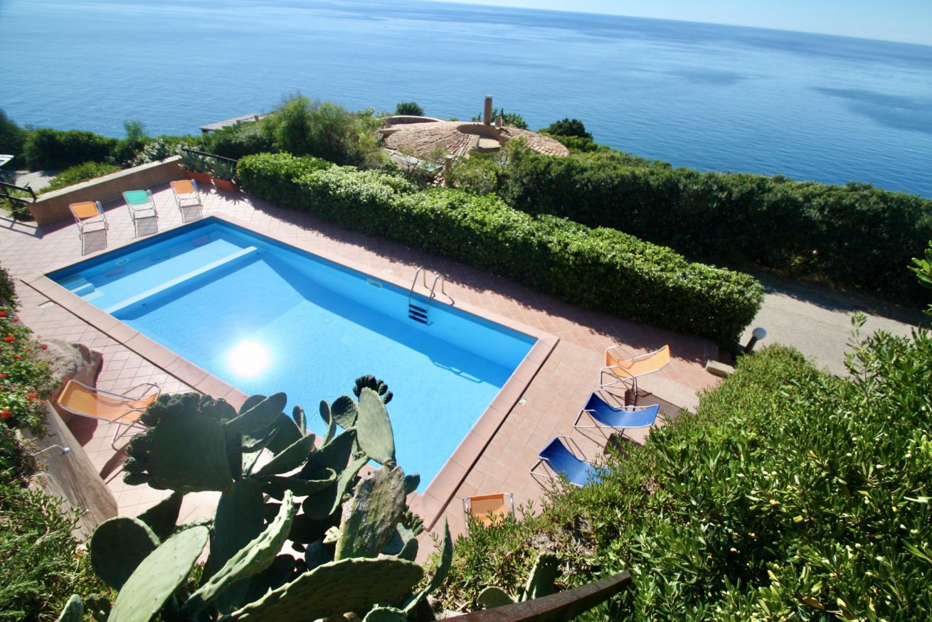 A rectangular pool with a clear blue water surface, surrounded by a low brick wall. There are several lounge chairs and a small table near the pool. The pool is set against a backdrop of a clear blue sky and a body of water.