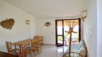Un coin repas avec une table et des chaises en bois, un canapé agrémenté de coussins à motifs et une porte-fenêtre coulissante donnant sur une terrasse extérieure.