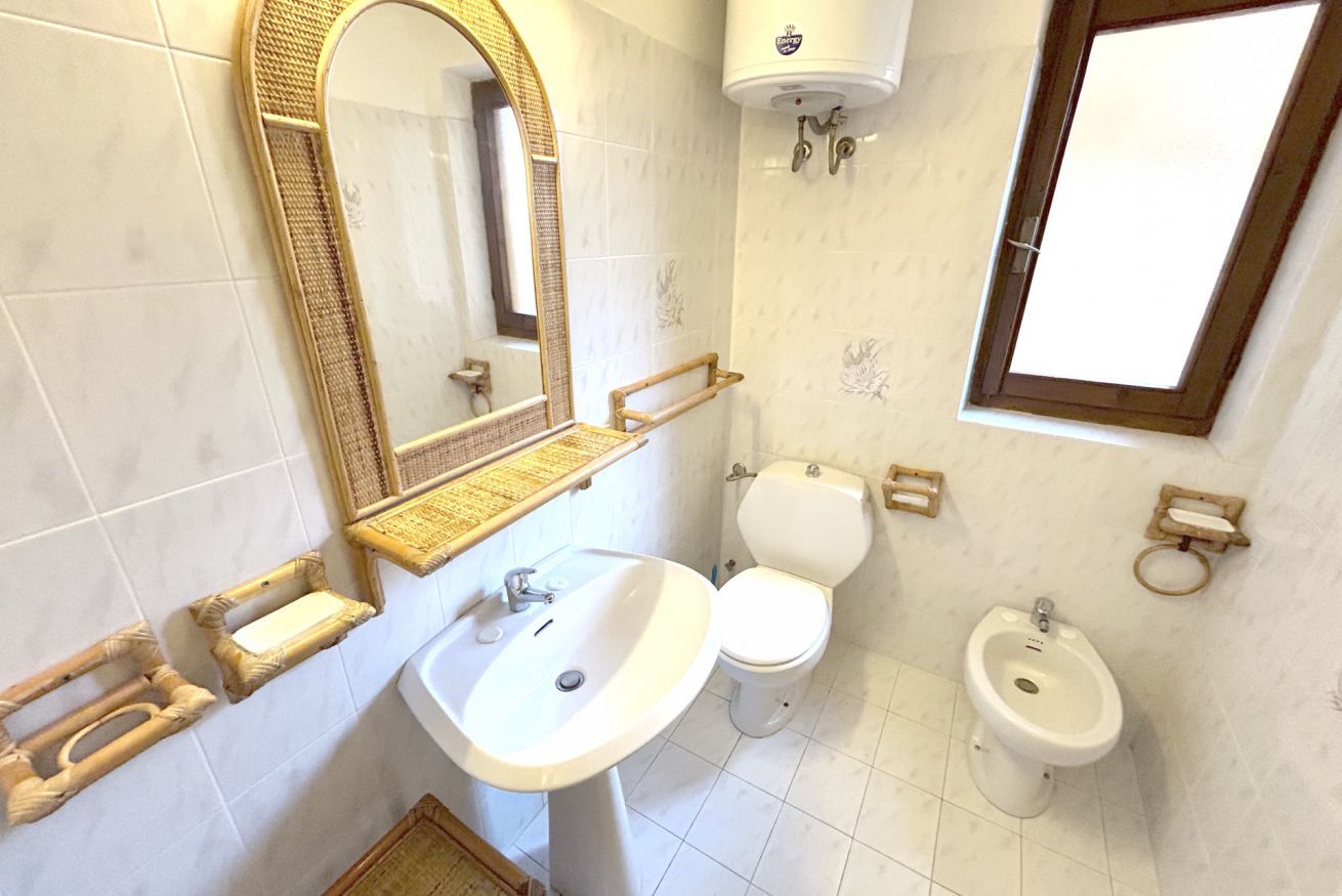 A bathroom with a white sink, two toilets, a bidet, a mirror with a wooden frame, and a window.
