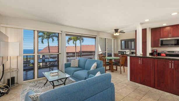 A spacious living area with a blue sofa, a dining table, and a kitchen with red cabinets and a microwave. - 🌴Beautiful condo located at higher elevations, Expansive Ocean Views🌴 (Holualoa)