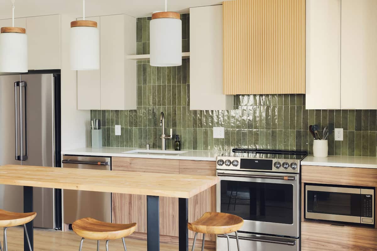 A modern kitchen with a wooden table, bar stools, stainless steel appliances, and pendant lights.