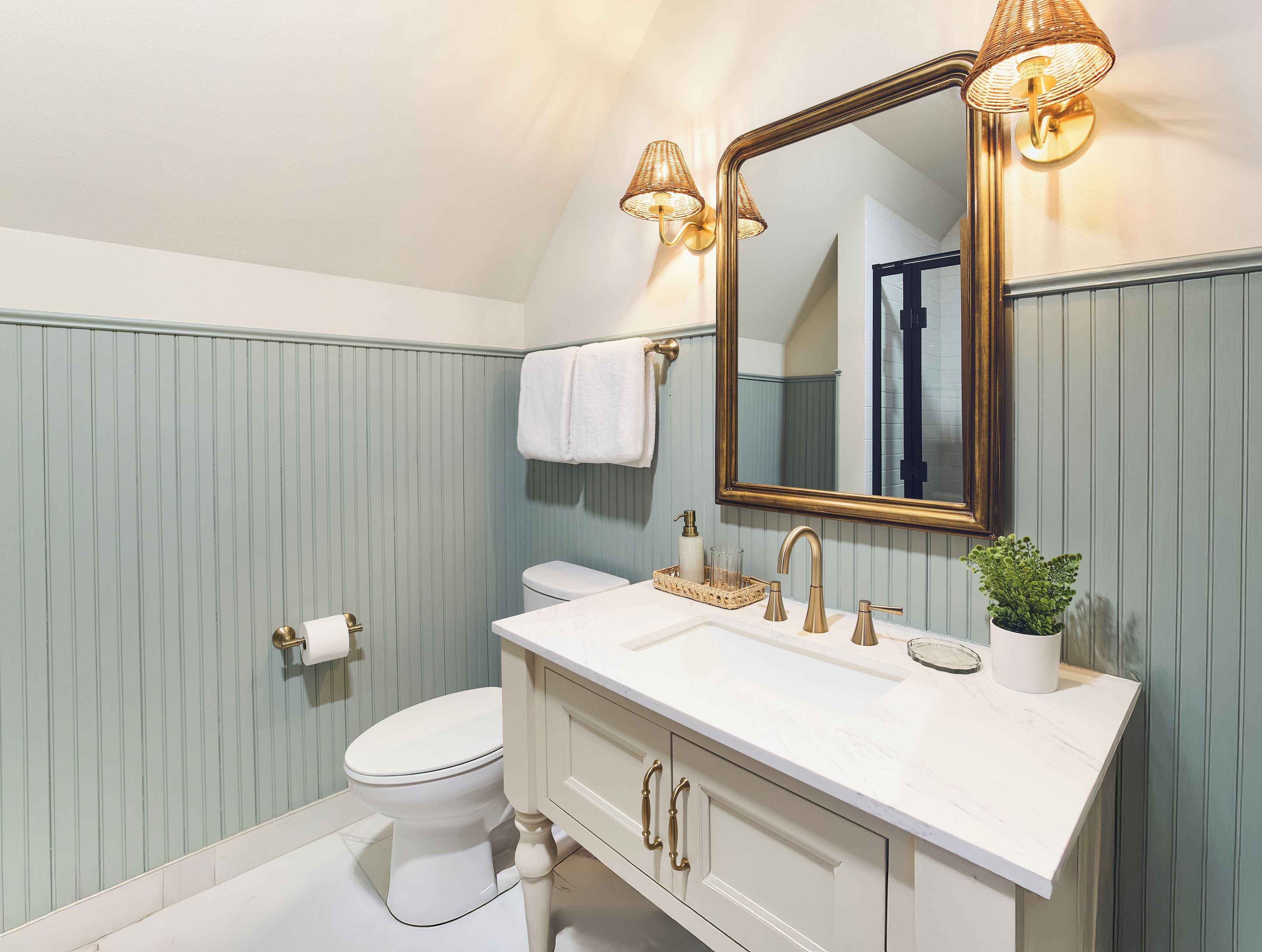 Une salle de bain avec un lavabo blanc, des robinetteries dorées, un grand miroir et une plante en pot.