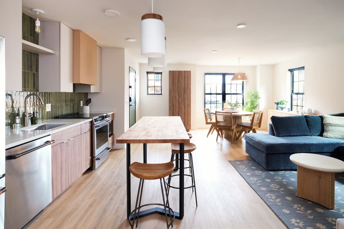 A modern kitchen with a wooden table and bar stools, a dining area with a table and chairs, and a blue sofa in a living room.
