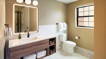 A bathroom with a white sink, wooden vanity, a large mirror with lights, a toilet, and a window.