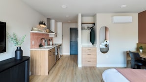 Une cuisine moderne avec des armoires en bois, un four encastré et un évier. On y trouve un coin repas avec une table et des chaises, ainsi qu’un lit recouvert d’une couverture rose.
