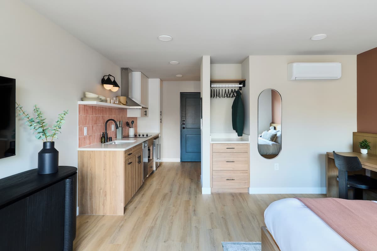 A modern kitchen with wooden cabinets, a built-in oven, and a sink. There is a dining area with a table and chairs, and a bed with a pink blanket.