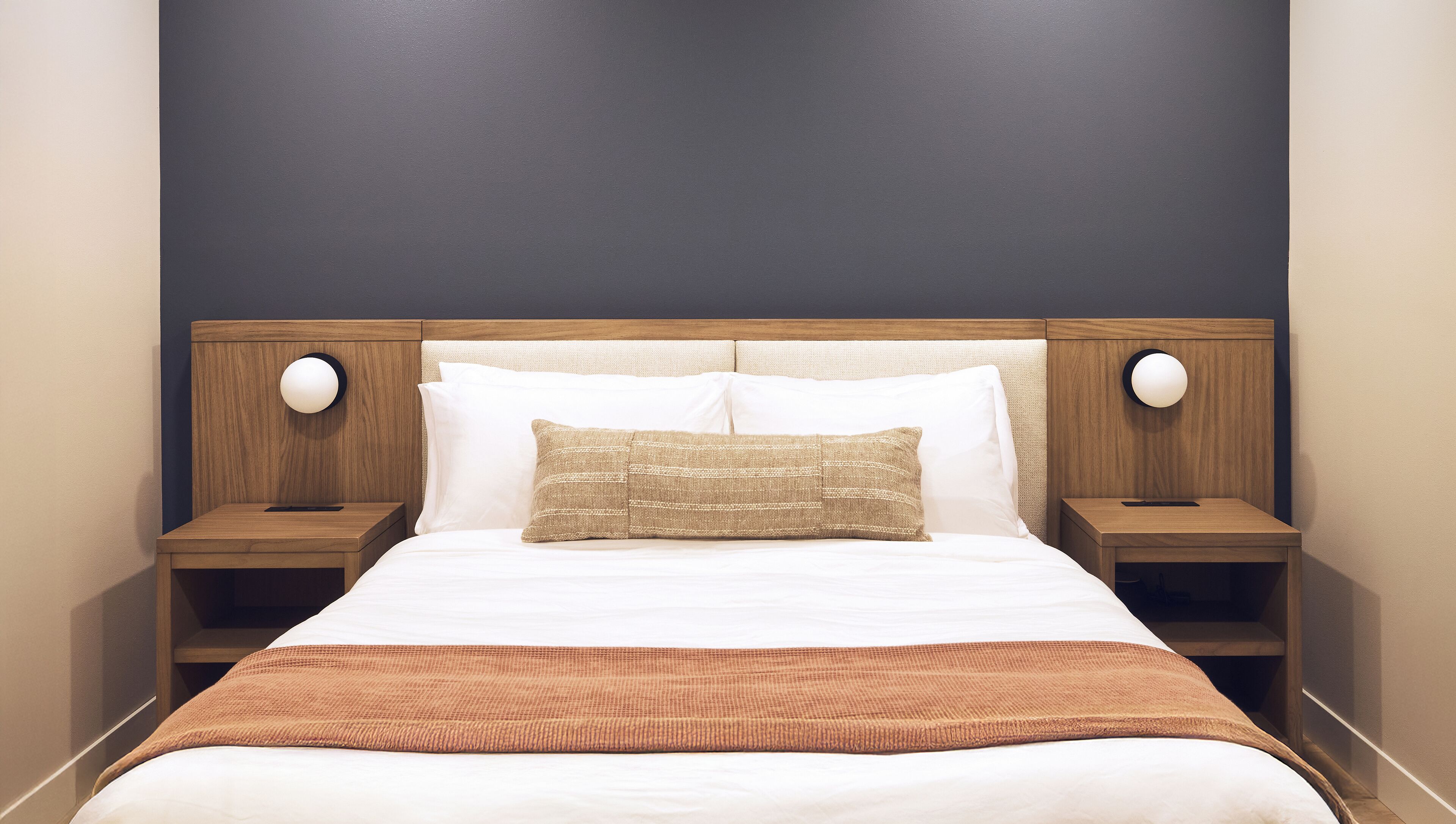 A neatly made bed with white and brown bedding, two bedside tables with lamps, and a dark wall behind the bed.