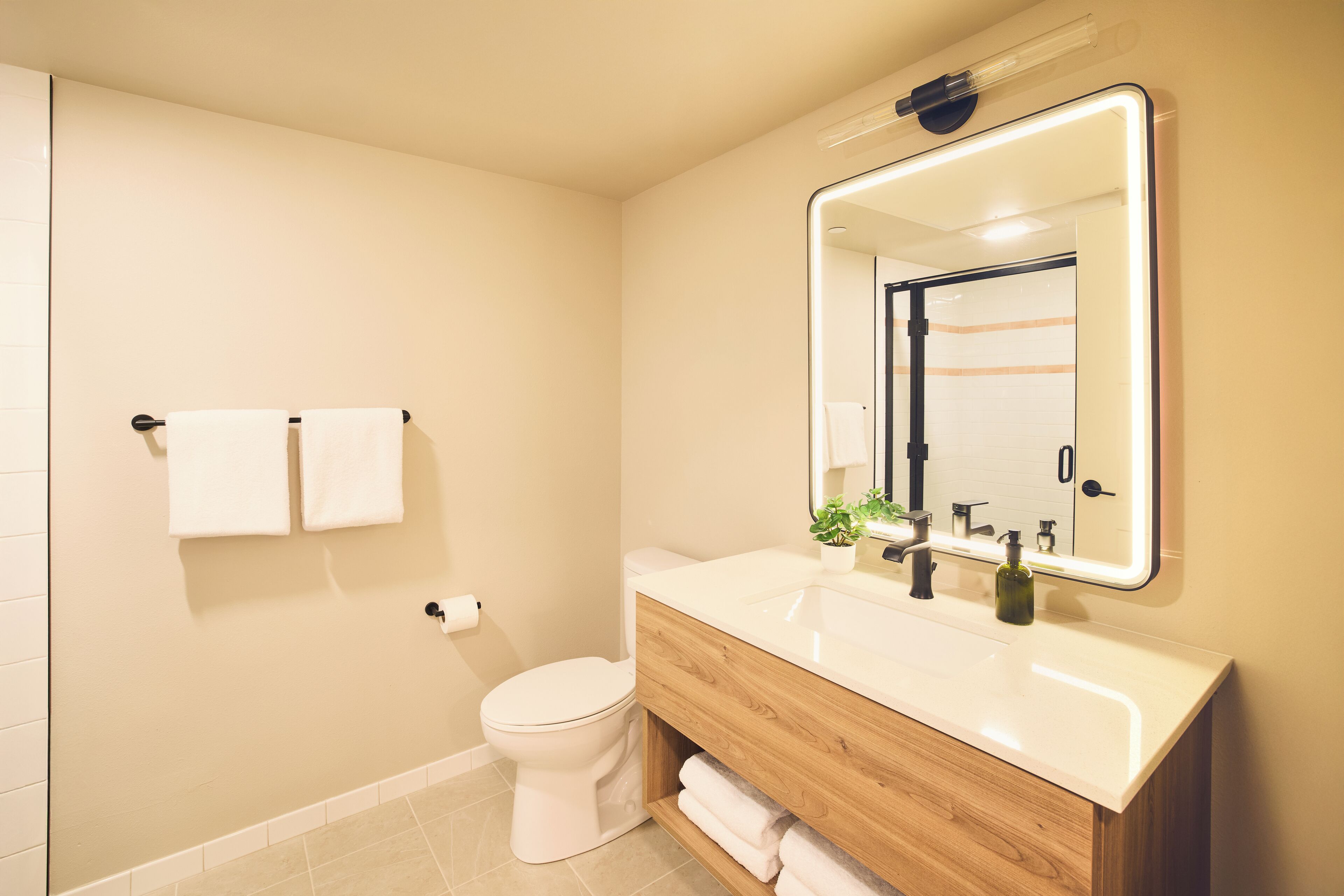 Une salle de bain moderne avec des toilettes blanches, un meuble-lavabo en bois, un grand miroir et deux serviettes blanches accrochées au mur.