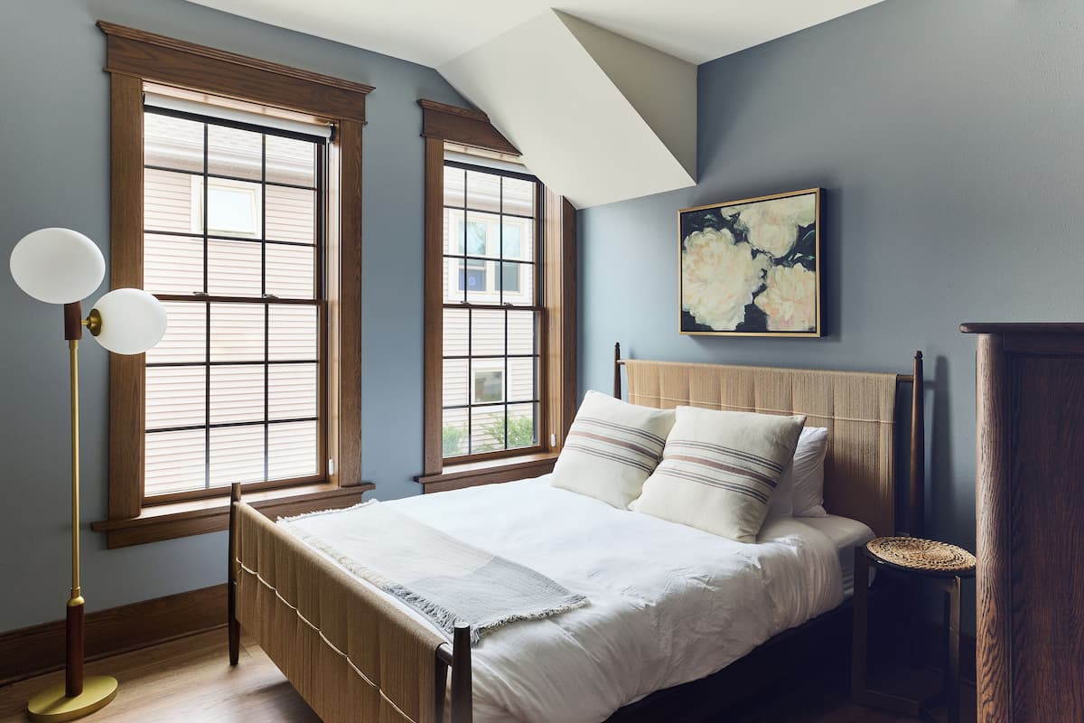 A bedroom with a bed, two windows, a floor lamp, a bedside table, and a painting on the wall.