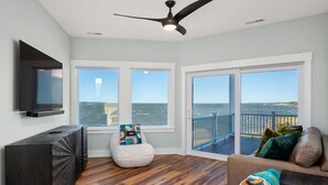 A spacious room with a large window overlooking the ocean, a ceiling fan, a flat-screen TV mounted on the wall, and a wooden floor. - Kite Surf Cottage: Stunning Soundfront Home, Panoramic views of the Sound and Sunset (Nags Head)