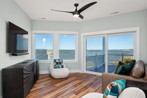 A spacious room with a large window overlooking the ocean, a ceiling fan, a flat-screen TV mounted on the wall, and a wooden floor. - Kite Surf Cottage: Stunning Soundfront Home, Panoramic views of the Sound and Sunset (Nags Head)