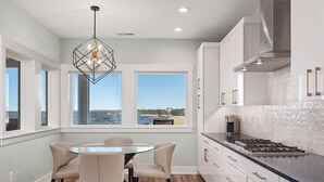 A modern kitchen with a gas stove, white cabinets, and a dining area with a round table and chairs. - Kite Surf Cottage: Stunning Soundfront Home, Panoramic views of the Sound and Sunset (Nags Head)
