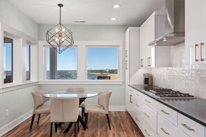 A modern kitchen with a gas stove, white cabinets, and a dining area with a round table and chairs. - Kite Surf Cottage: Stunning Soundfront Home, Panoramic views of the Sound and Sunset (Nags Head)
