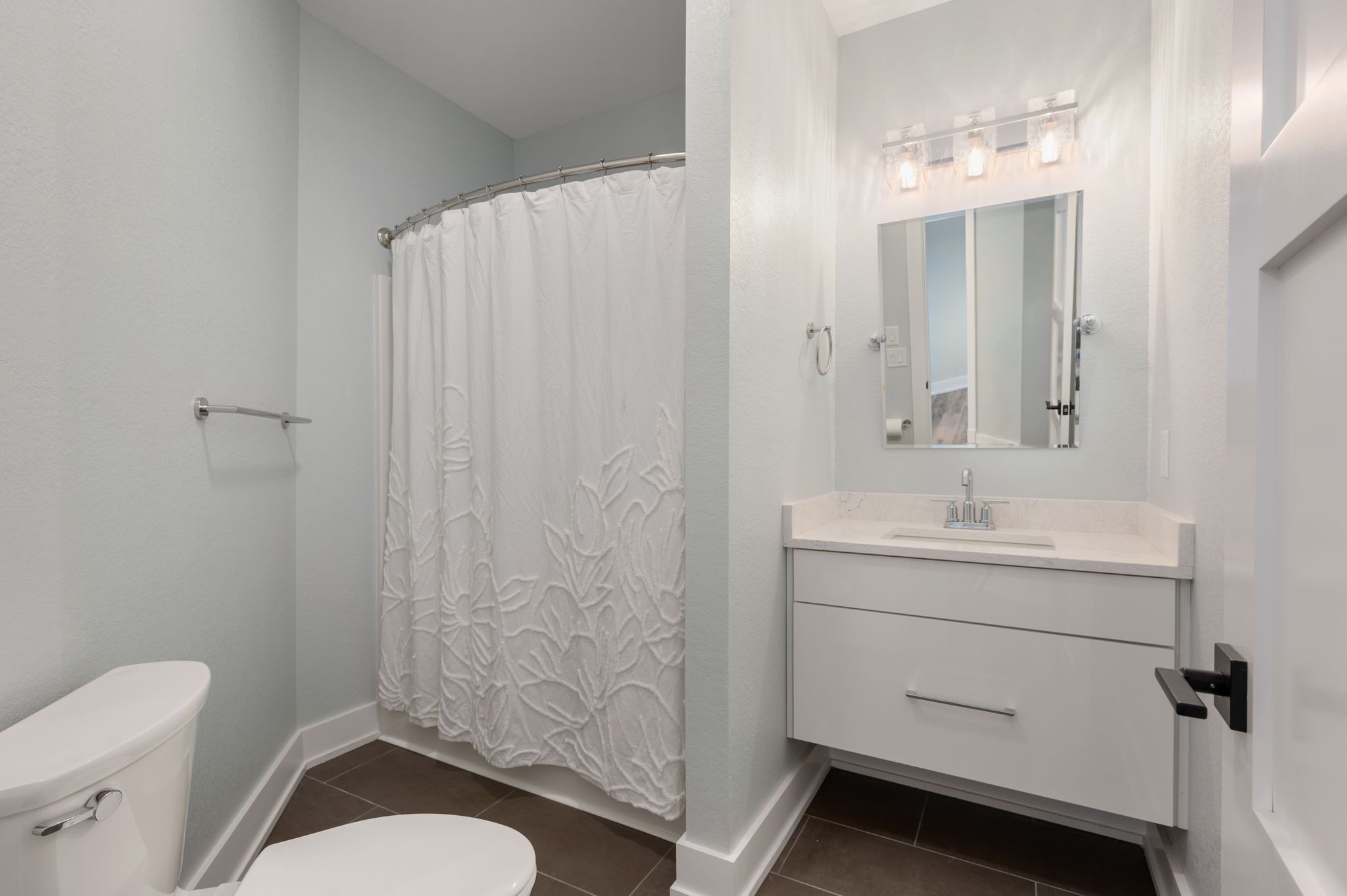 A bathroom with a toilet, shower curtain, vanity with a mirror and lights, and a sink.