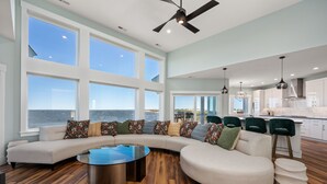 A modern living room with a curved sectional sofa, a glass coffee table, and a view of the ocean. - Kite Surf Cottage: Stunning Soundfront Home, Panoramic views of the Sound and Sunset (Nags Head)