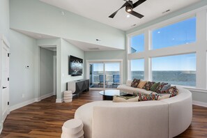 A spacious living room with a large window overlooking the ocean, a ceiling fan, and a modern seating area.