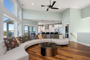 A spacious living room with a curved sectional sofa, a glass coffee table, and a ceiling fan. - Kite Surf Cottage: Stunning Soundfront Home, Panoramic views of the Sound and Sunset (Nags Head)