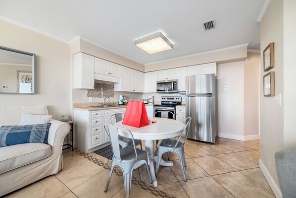 A compact kitchen with white cabinets, a stainless steel refrigerator, and a microwave. There is a dining table with two chairs and a red bag on it.