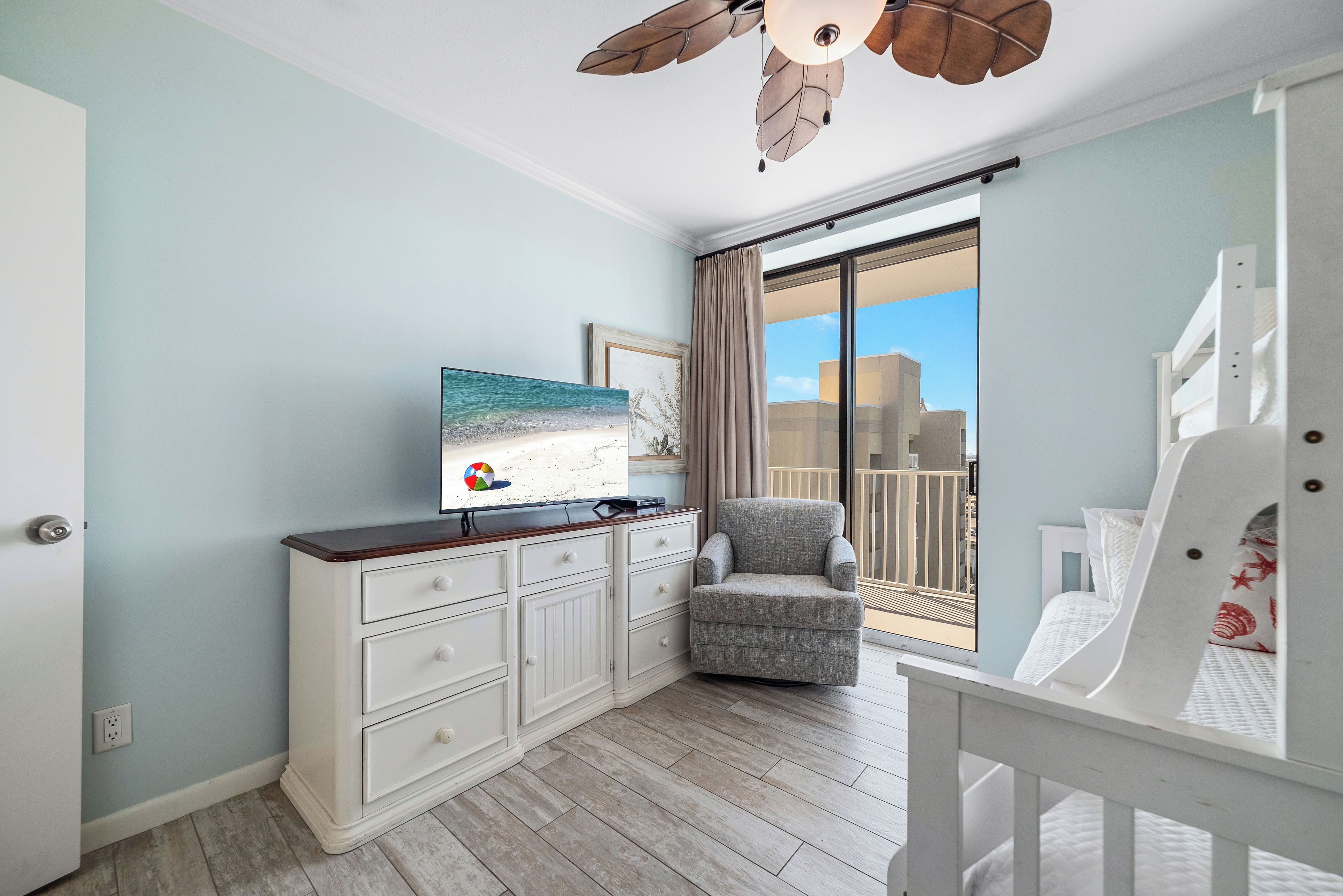 A room with a white dresser, a gray armchair, a bed with white bedding, a ceiling fan, and a large window with a view of buildings.