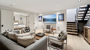 A modern living room with a sofa, armchairs, a coffee table, and a TV. There is a staircase leading to a loft area.