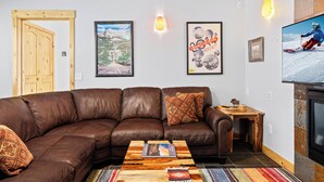 A modern living room with a brown leather sectional sofa, a wooden coffee table, and a flat-screen TV mounted on the wall.