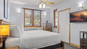 A bedroom with a bed, a ceiling fan, a wall-mounted TV, and a window with blinds.