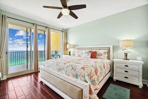 A bedroom with a bed, a nightstand, a lamp, a ceiling fan, and a view of the ocean through a sliding door.