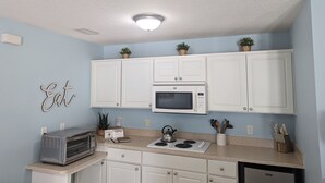 A kitchen with white cabinets, a microwave, a toaster, and a stove. There are potted plants on the cabinets and a sign that says "Eat". - Charming Cocoa Village Hideaway (Cocoa)