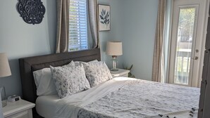 A bedroom with a bed, nightstands, a lamp, a decorative wall piece, and a window with curtains. - Charming Cocoa Village Hideaway (Cocoa)