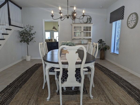 A dining room with a round table, white chairs, a chandelier, a clock, and a window with blinds.