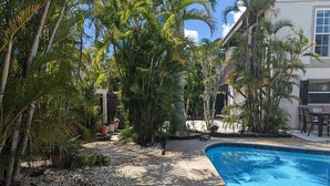 A pool area with a concrete deck, surrounded by palm trees and a white building. - Charming Cocoa Village Oasis (Cocoa)