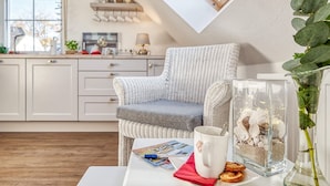 A cozy kitchen with a high ceiling, a wicker chair, a coffee table with magazines, a mug, and a decorative glass container with seashells.