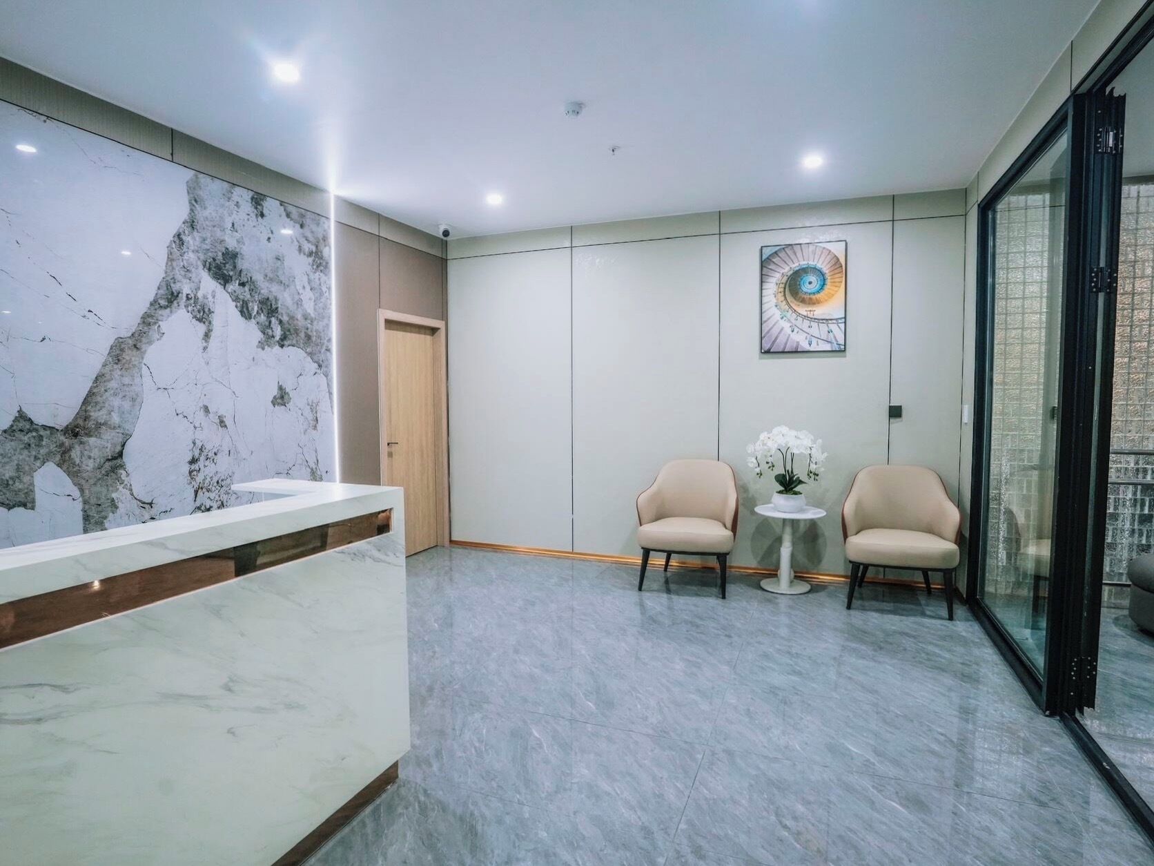 A modern reception area with a marble wall, two seating chairs, a small table with flowers, and a glass partition.