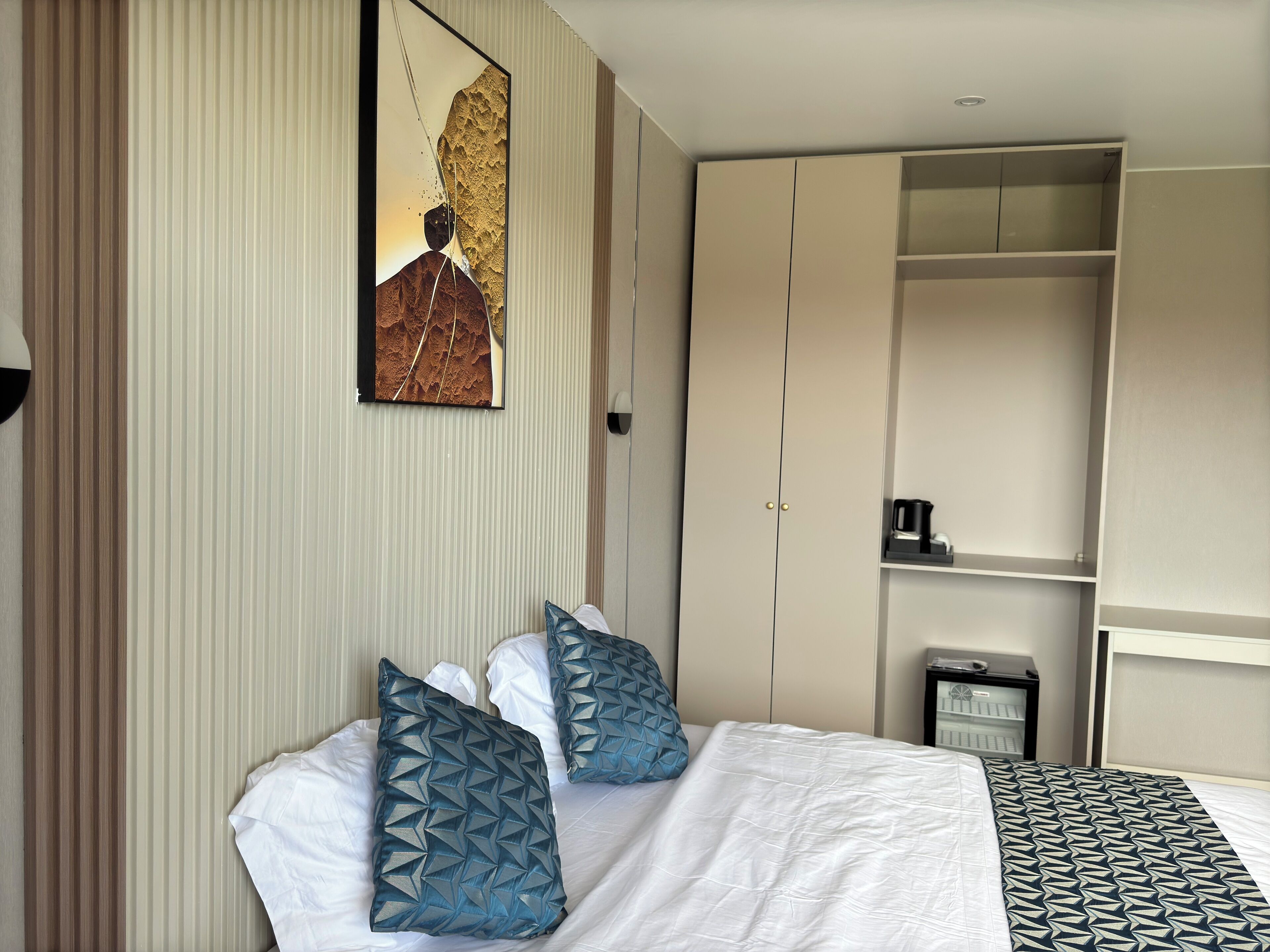 A hotel room with a bed, two pillows, a nightstand, a coffee maker, and a wardrobe.