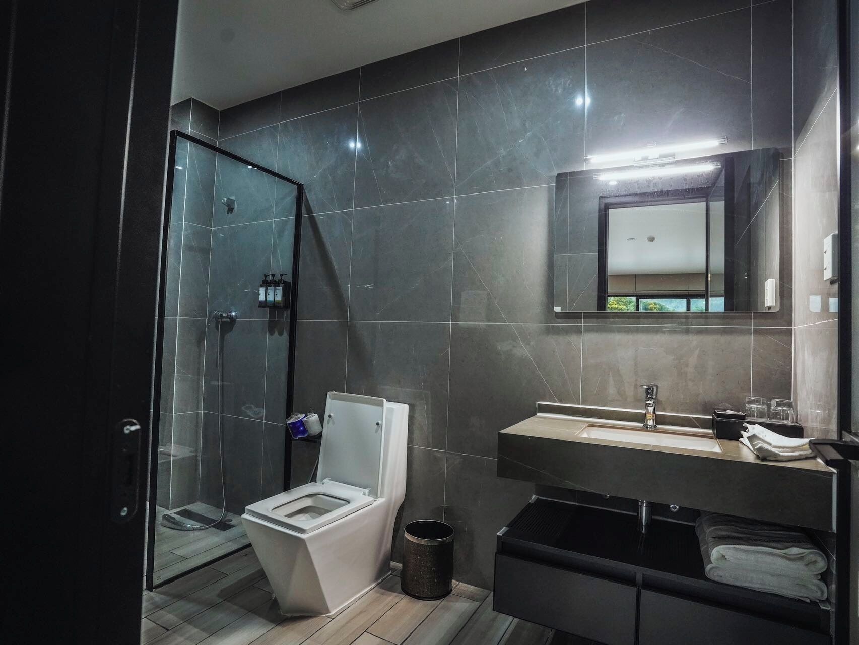 A modern bathroom with a glass shower enclosure, a white toilet, a sink with a mirror, and a trash bin.