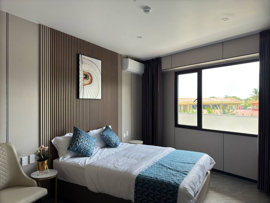 Una habitación de hotel moderna con una cama grande, una mesita redonda pequeña, una silla y un ventanal con vistas a edificios y vegetación.