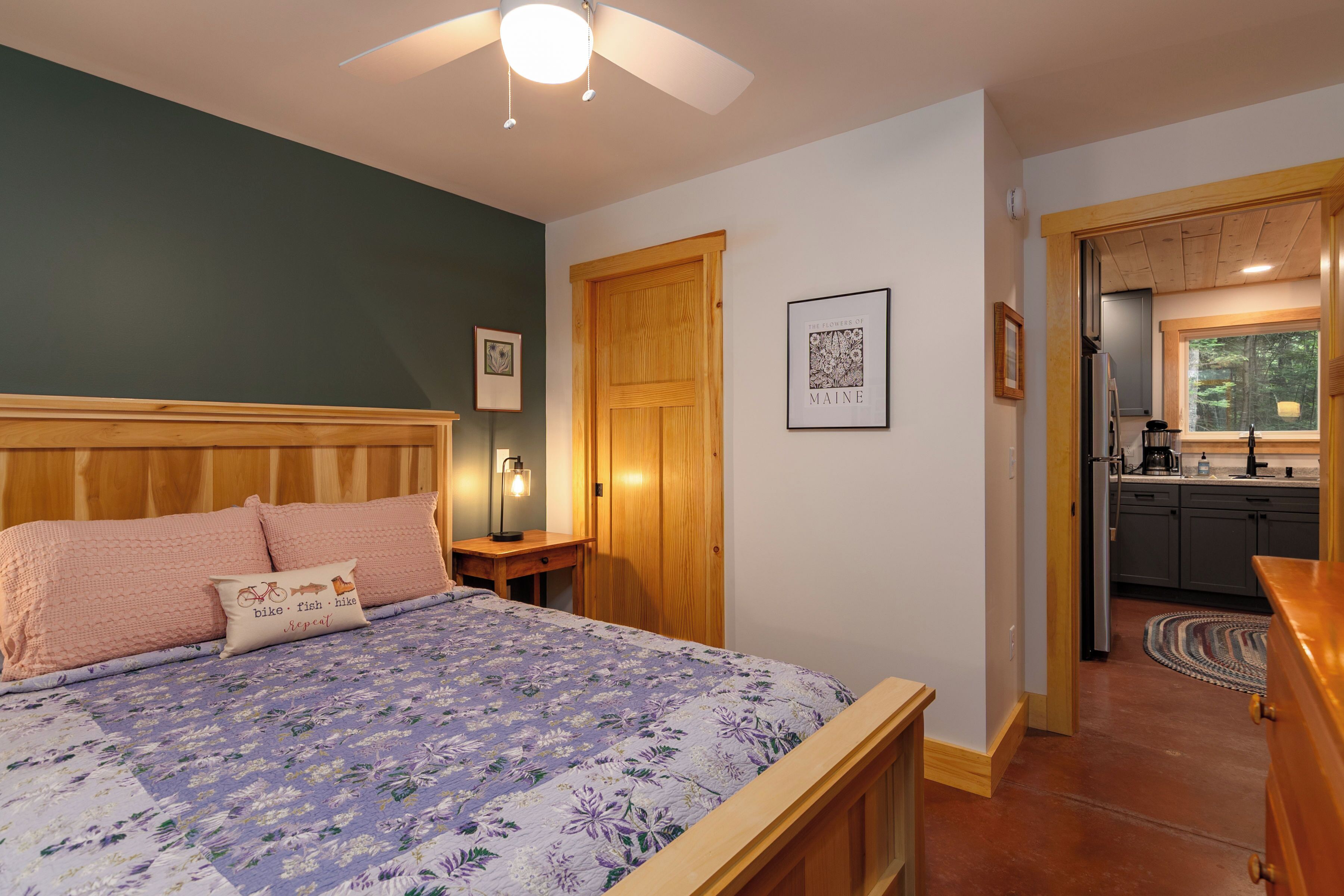 A bedroom with a bed, wooden headboard, a nightstand, a lamp, a framed picture, and a door leading to a kitchen.