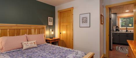 A bedroom with a bed, wooden headboard, a nightstand, a lamp, a framed picture, and a door leading to a kitchen.