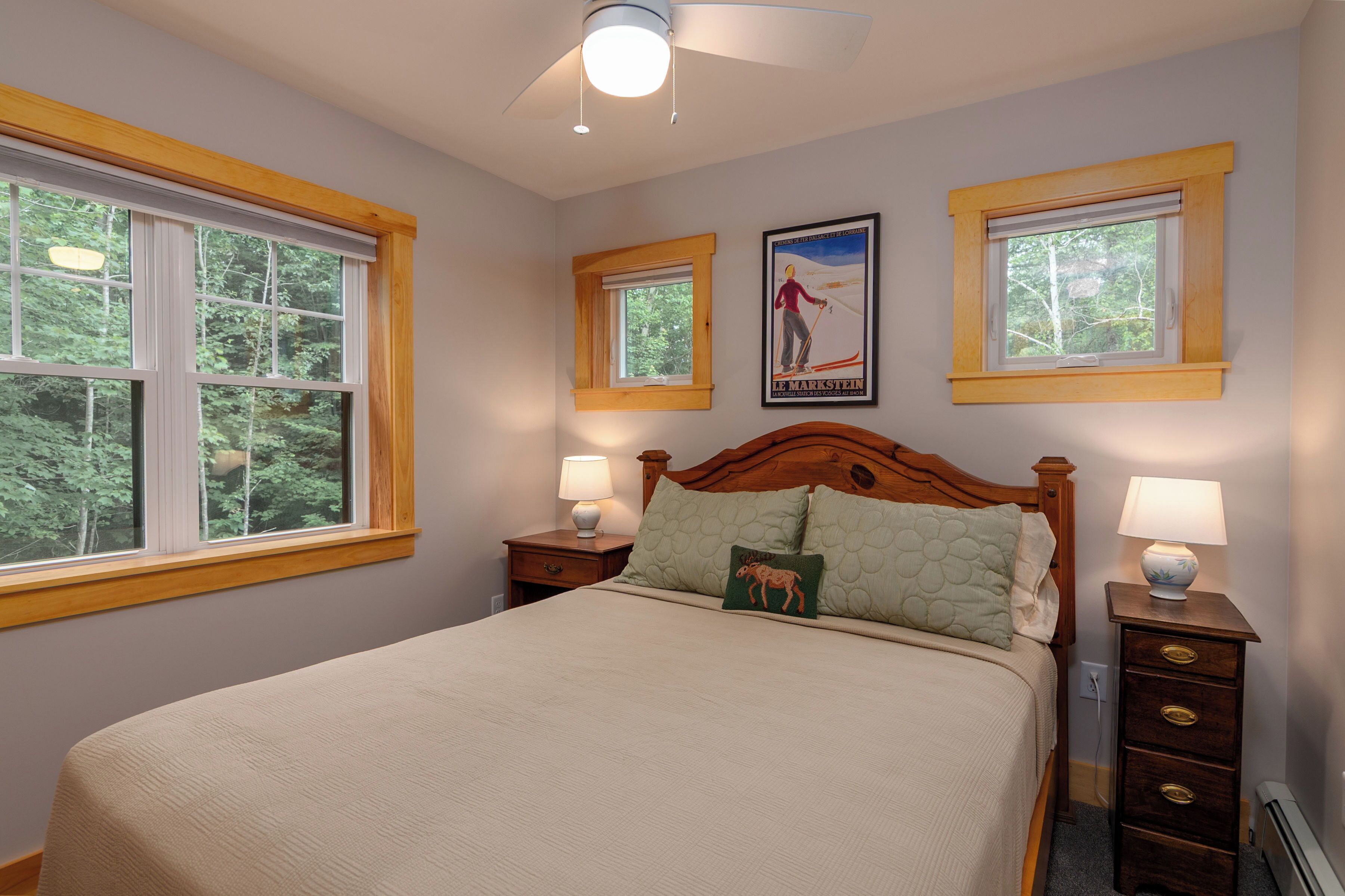 A bedroom with a large bed, two bedside tables with lamps, a headboard, and two windows with wooden frames.