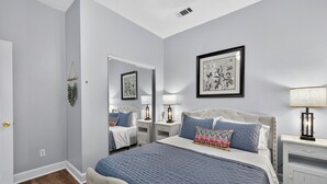 Une chambre à coucher avec un grand lit, deux tables de chevet, une commode, un miroir et une œuvre d’art encadrée accrochée au mur.