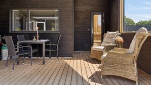 Una terraza de madera con una mesa y sillas, un sofá de mimbre y un vidrio que da a un espacio interior.