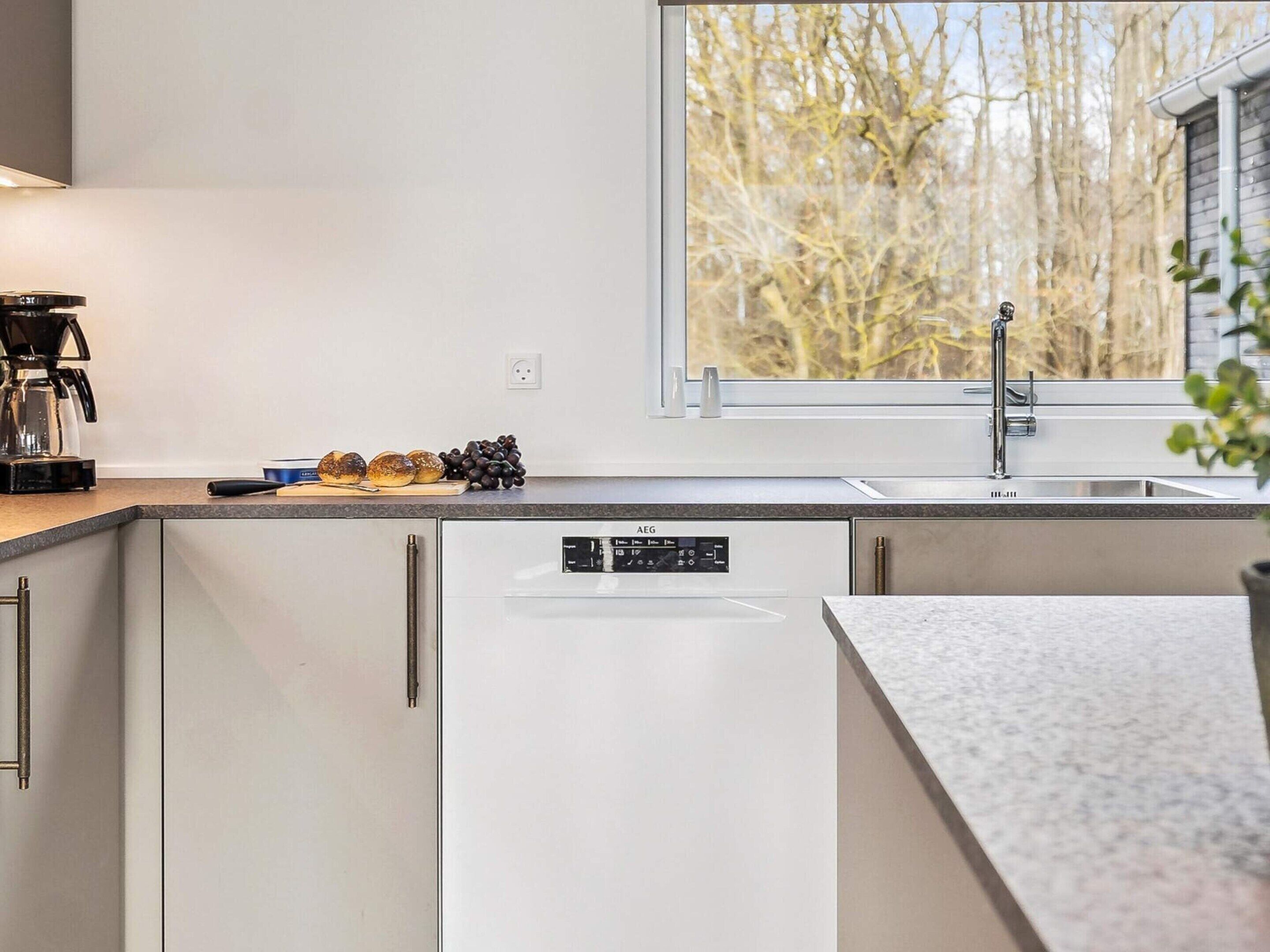 A modern kitchen with a built-in dishwasher, a window with a view of trees, and a countertop with a coffee maker and bread.