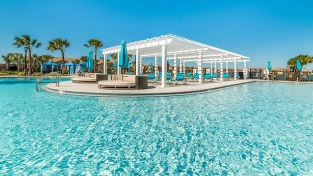 Una piscina azul cristalina con una pérgola blanca y sillas de descanso, rodeada de palmeras y sombrillas.