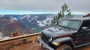 Een zwarte Jeep met de tekst 'MOJAVE' en 'Jeep' erop, geparkeerd op een onverhard pad met uitzicht op een kloof en wolken erboven.