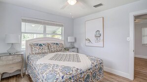 A bedroom with a large bed, two bedside tables, a window with blinds, a ceiling fan, and a framed picture on the wall. - Charming cottage in delightful Stuart with AC, WiFi (Stuart)