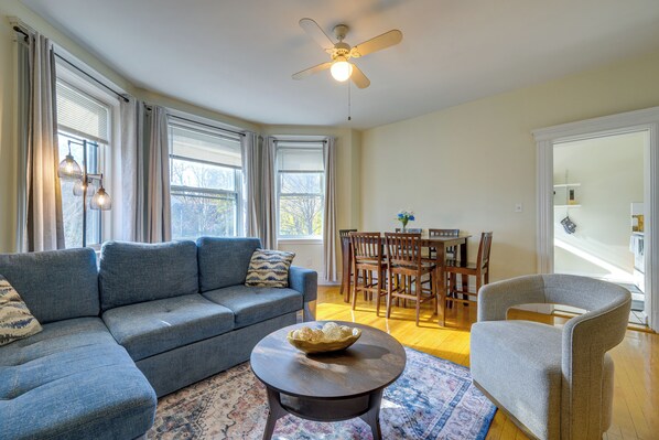 A living room with a blue sofa, a round coffee table, a gray armchair, and a dining area with wooden chairs and a table.