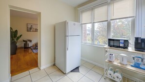 Une cuisine compacte équipée d’un réfrigérateur, d’un four à micro-ondes et d’une machine à café sur un plan de travail, d’une fenêtre avec des stores et d’une porte donnant sur une autre pièce.