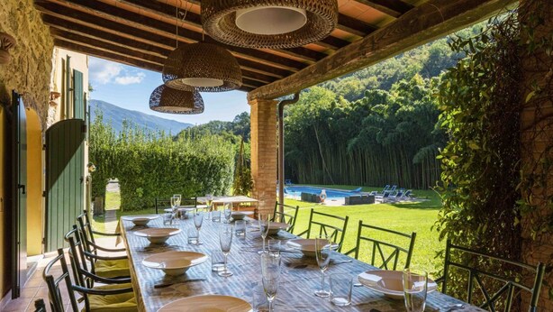 Una zona d'esmorzar a l'aire lliure amb una taula preparada per a sis persones, envoltada de cadires, i amb vista a un jardí exuberant i muntanyes al fons.